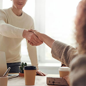  Two people shake hands across a table, with a third person partially visible.