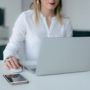 Woman in a white shirt using a computer mouse next to a laptop and a smartphone, symbolizing office work.