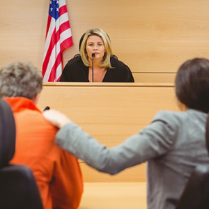 A blonde female judge or witness speaking into a microphone in a courtroom with the American flag in the background.