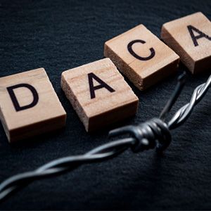 Wooden blocks spelling out "DACA" resting on a dark surface next to a barbed wire fence, symbolizing immigration policy and protection.